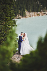 Beautiful wedding photo on mountain lake. Happy Asian couple in love, bride in white dress and groom in suit are photographed against background of the Kazakh landscape