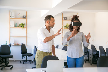 Young woman in VR glasses standing in office, touching air. Her male colleague instructing her, showing hand gestures. Virtual reality experience concept