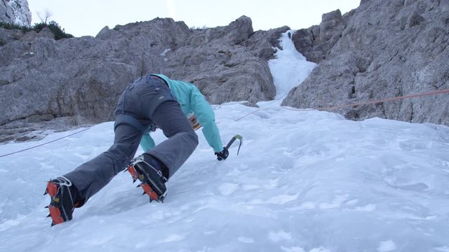 SLOW MOTION, BOTTOM UP, CLOSE UP: Woman ice climbing stops and looks up at beautiful frozen waterfall. Fearless female tourist goes ice climbing up the gorgeous icy stream running down the rocky cliff