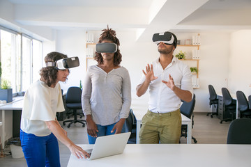 Diverse team of three watching virtual presentation. Man and women wearing virtual reality glasses, standing in office, using laptop, touching air. Teamwork concept