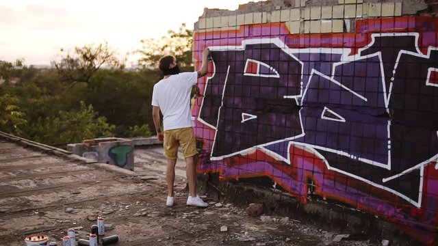 A young man with a spray can finishing draw a new graffiti on the wall. Video of the process of drawing a graffiti on a bricked wall on the roof. Slow motion paint application the white lines in word