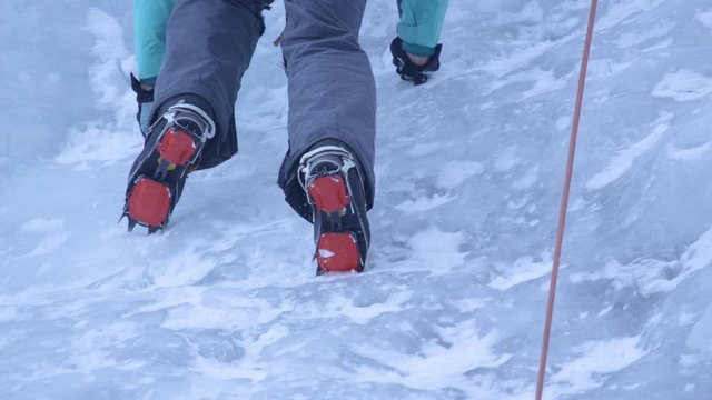 SLOW MOTION, CLOSE UP, BOTTOM UP: Cinematic shot of climbers crampons being kicked into the icy river cascading down a cliff. Woman wearing crampons over her hiking boots climbs up a frozen waterfall