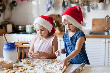Children are cooking Christmas cookies in cozy home kitchen. Cute kids prepare holiday food for family. Funny little sisters in flour make pastries. Lifestyle candid moment. Children chef concept