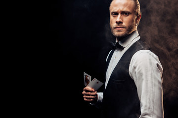 handsome croupier holding playing cards on black with smoke