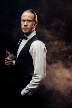 handsome croupier holding playing cards on black with smoke