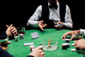 cropped view of men near croupier in formal wear with playing cards isolated on black