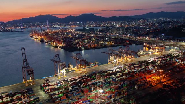 Timelapse illuminated cranes and containers on Busan cargo port unloading site at sea surface reflecting sunlight against hill silhouettes under orange sunset sky zoom in