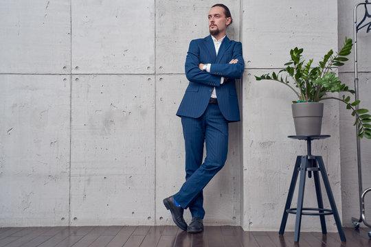 Full height image of brunet man in business suit next to houseplant on gray concrete wall.