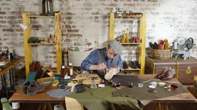 Professional male shoemaker dyeing new leather boot with brush while working at desk in studio