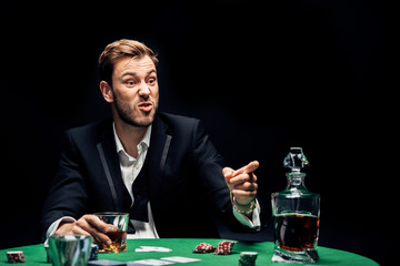 selective focus of angry man pointing with finger near alcohol and playing cards isolated on black