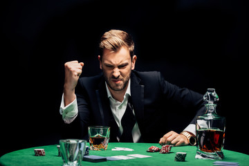 selective focus of angry man with clenched hand near alcohol and playing cards isolated on black