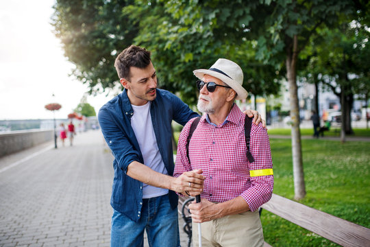 Young man and blind senior with white cane walking on pavement in city.