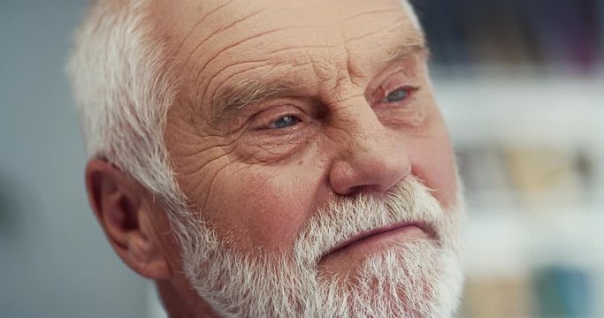 Close up of the face of the old Caucasian grey-haired man with a beard looking at the side with thoughtful face. Indoor.