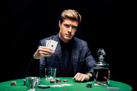 selective focus of handsome man holding playing cards near poker chips and alcohol drink isolated on black
