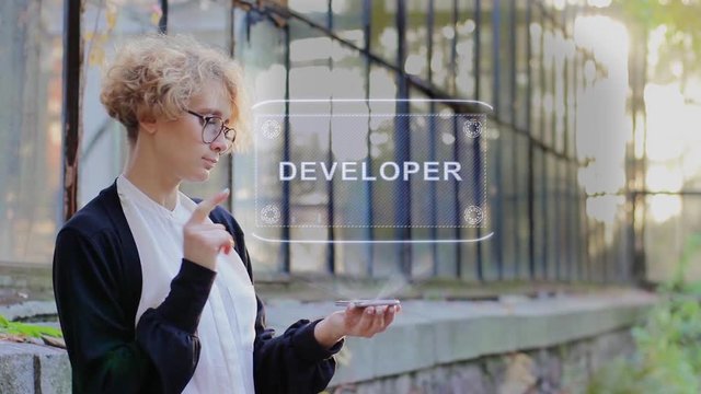 Curly young woman in glasses interacts with a hud hologram with text Developer. Blonde girl in white and black clothes uses technology of the future mobile screen