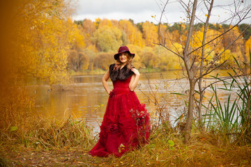 Young woman in stylish hat and long red evening dress with fur posing near the river and walking in the autumn garden background alone