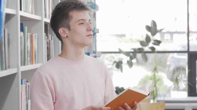 Young man enjoying reading a book at the library. Charming male student looking away dreamily while reading a fascinating book at bookstore. Inspiration, imagination concept