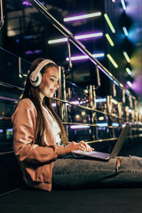 smiling woman in pink jacket with headphones using laptop in night city