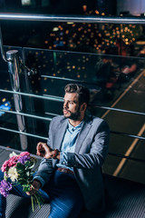 handsome businessman in formal wear with bouquet looking away in night city