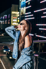 Naklejka na ściany i meble attractive woman in denim jacket and sunglasses smiling and looking away in night city