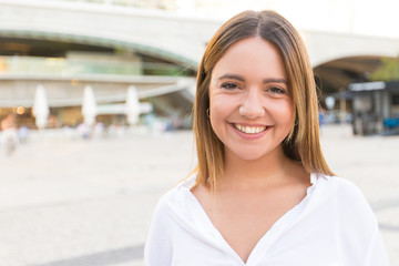 Happy carefree lady posing in city square. Front portrait on fair haired young woman in white casual shirt looking and smiling at camera. Female portrait concept