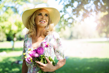 Hübsche ältere Frau im Sommerkleid im Park mit Blumen und Strohhut 