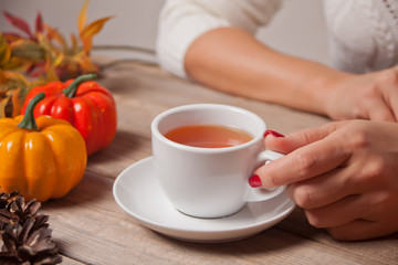 Woman's hand hold cup of tea. Autumn leaves, candle, pumpkin on the wooden background.