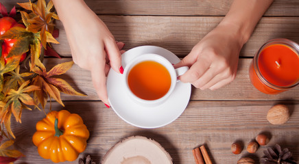 Woman's hand holding cup of tea. Autumn leaves, candle, pumpkin on the wooden background.