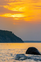 picturesque coast of the sea with a cliff in the background, large boulder in the water, low sun during sunset in orange light