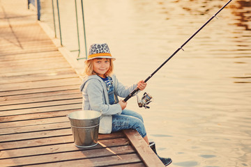 Cute child girl fishing from wooden pier on a lake. Family leisure activity during summer sunny day. little girl having fun by a river at beautiful summer evening. Active family time on nature