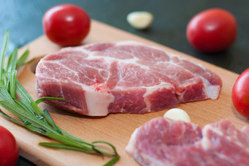 Raw juicy pork steaks with red tomatoes, rosemary and garlic on a cutting board.