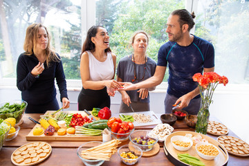 Happy friends preparing healthy meal. Cheerful young man and women in sportswear standing at table with raw organic fruits and vegetables. Healthy eating concept