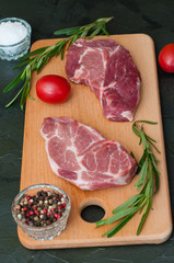Raw meat steaks, tomatoes, rosemary, salt and pepper on dark background, vertical frame