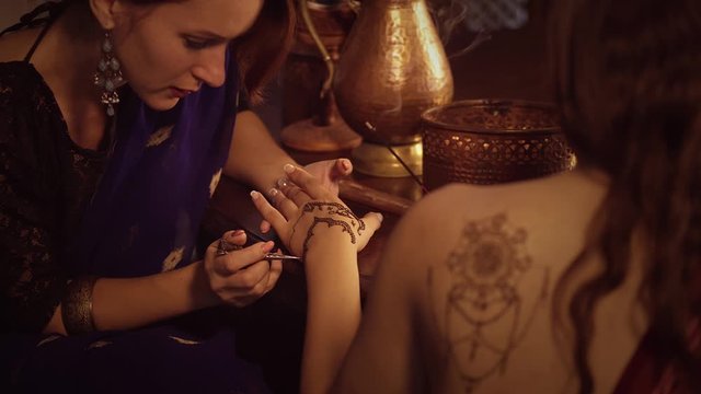 Master is performing mehndi beauty treatments in studio. Woman is decorating hands of client girl by temporary henna tattoos