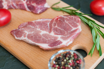 Raw pieces of pork, pepper, rosemary and tomatoes on cutting board, on a dark gray concrete background, close-up