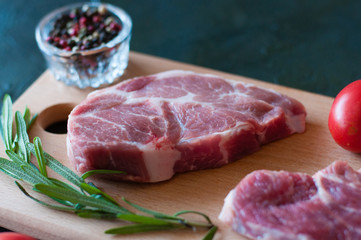 Raw pork steak, rosemary, pepper and tomato on a cutting board, on dark gray concrete background, close up, in dark tonality