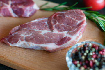 Raw pork steak, rosemary, pepper and tomato on a cutting board, on dark gray concrete background, close up, in dark tonality