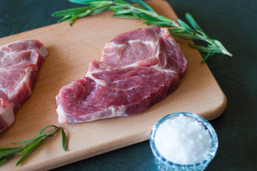 Raw pork steak, salt, rosemary and tomato on wooden cutting board, on a dark gray concrete background, close up, in dark tonality