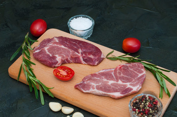 Two raw pork steaks, sea salt, rosemary, garlic, red tomatoes and peppers on a light wooden cutting board, on dark gray concrete background, medium plan, top view