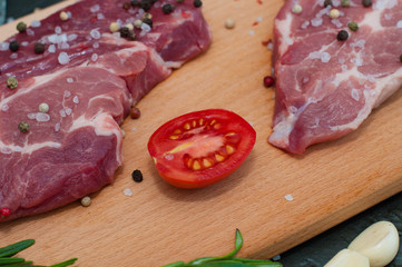 Raw pork meat on cutting board, sprinkled with salt and pepper, garlic, rosemary and tomato, top and side view
