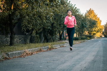 Fototapeta premium Girl runner running on sunset in autumn forest and park wearing pink jacket. Young woman jogging in fall colors. Sport and activities concept.