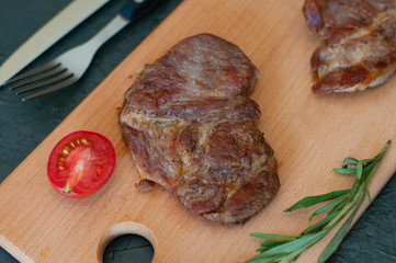 Grilled veal, beef or pork steak, tomato, rosemary and soy sauce for meat, knife and fork, on a wooden board, close-up, flat lay