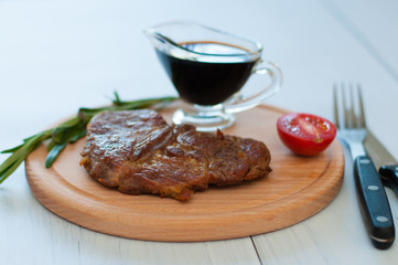 Cooked, grilled pork steak on a wooden round board, with sauce, rosemary and tomato, fork and knife, on a light background with copy space