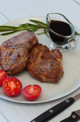 Tasty, grilled pork steaks on a gray plate with sauce, rosemary and tomatoes, on a light wooden background, cutlery, vertical frame format