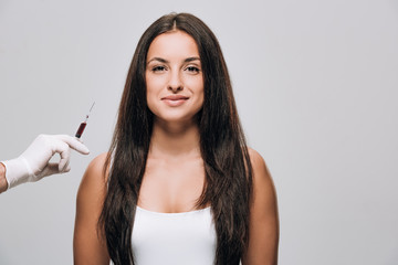 cosmetologist in latex gloves holding syringe for scalp mesotherapy near brunette beautiful woman...