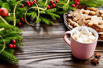 Christmas background of pink mug with hot chocolate and marshmallows, spruce branch and tray with gingerbread man cookies on wooden table