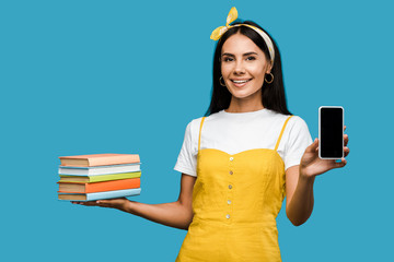 positive woman holding books and smartphone with blank screen isolated on blue