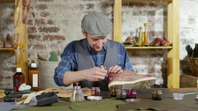 Zoom in of happy male shoemaker dyeing leather shoe and then looking at camera and smiling while working at desk in workshop
