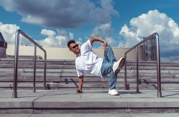 Male athlete, guy dancer in summer in city. Dancing in motion, break dance, fitness outdoor sports. In a T-shirt, leggings and sneakers. Background concrete stair clouds.