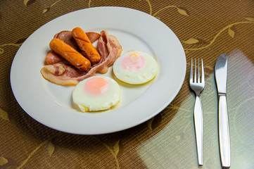 Yummy American breakfast with sunny fried eggs, rosted bacon, ham, two sausages in white dish with silver fork and knife on table.